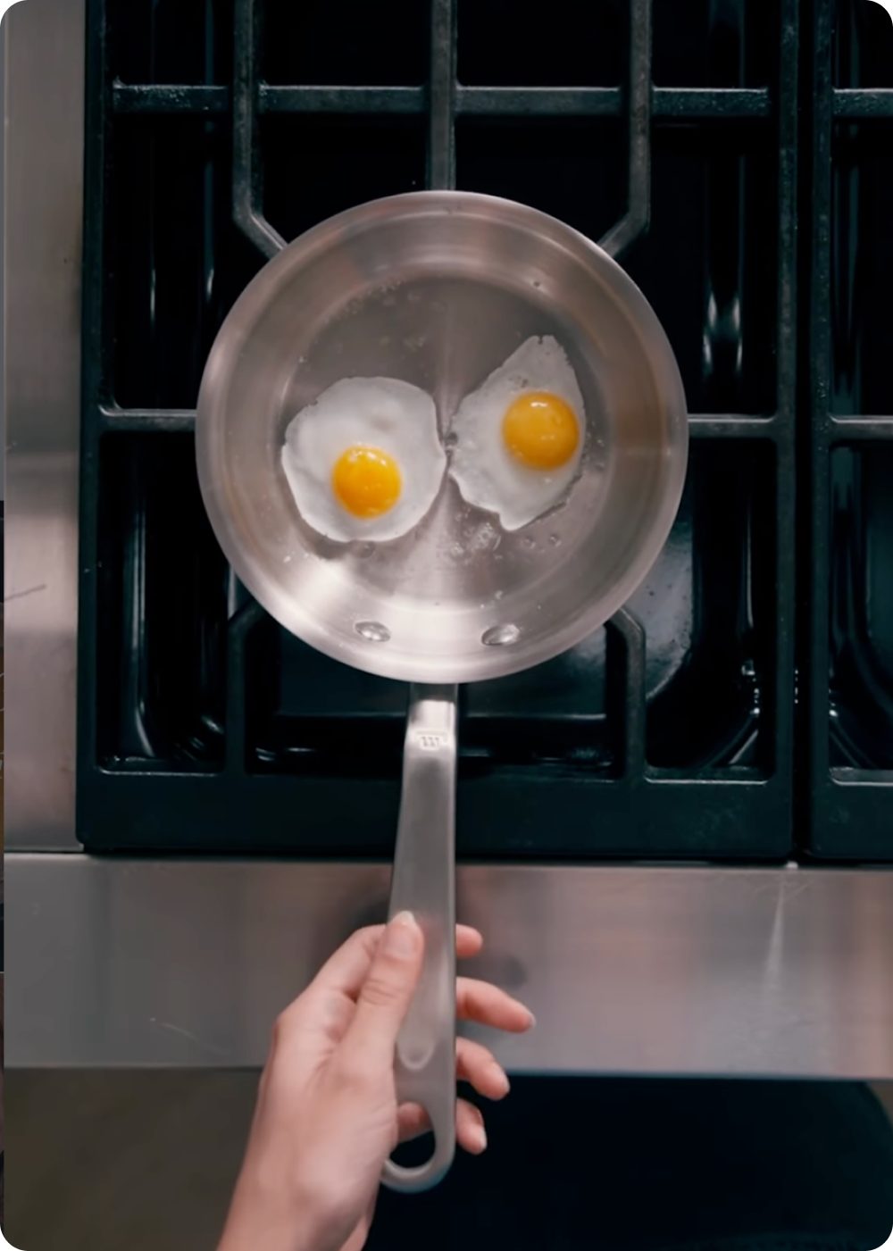Two eggs frying in a small stainless steel pan on a gas stovetop, held by hand