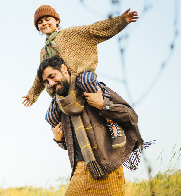 A smiling child in a brown knit sweater rides on his father’s shoulders as he walks leisurely through the tall grass. The child is wearing the Tejo from Wildling.