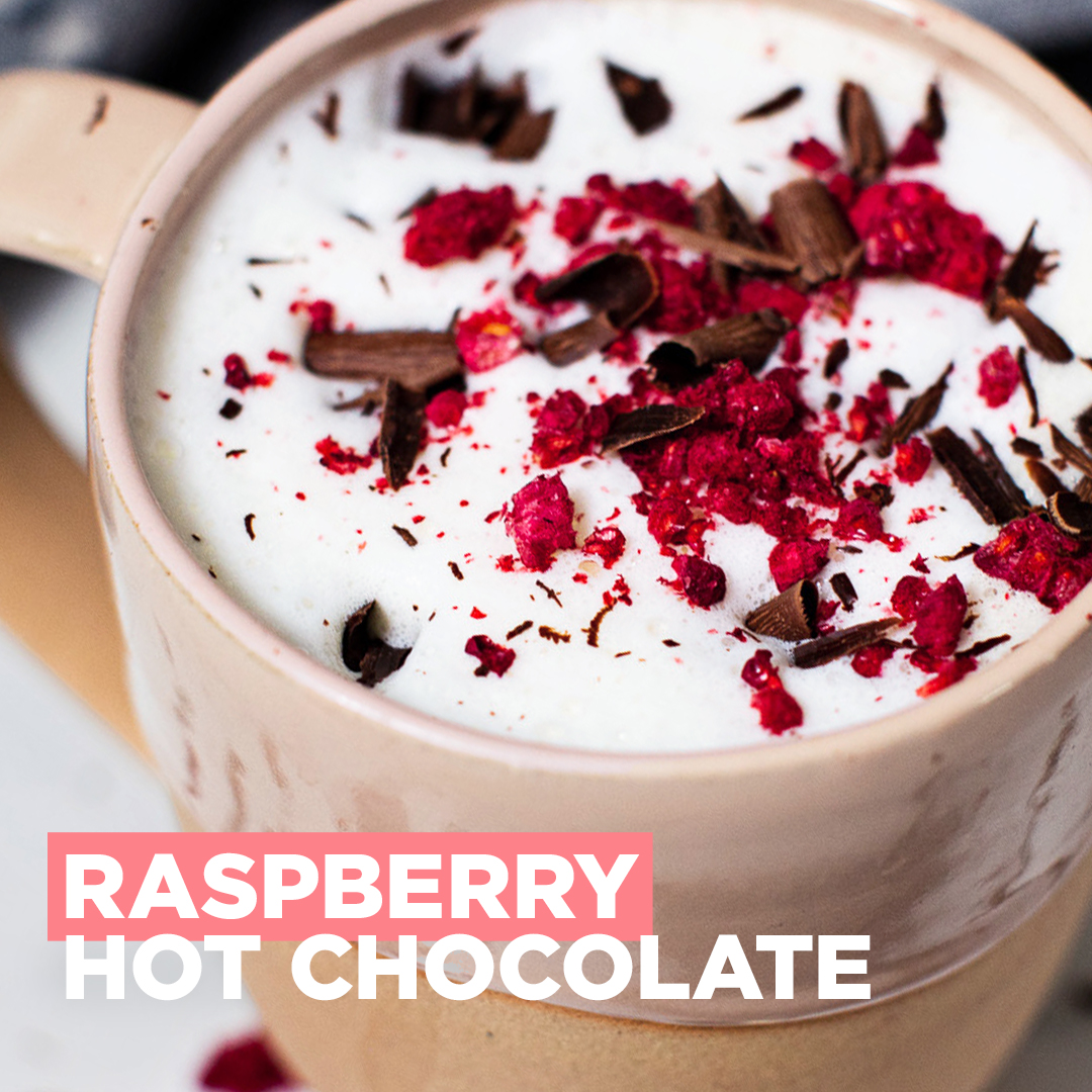 "Raspberry Hot Chocolate" showing a close-up of a ceramic mug topped with white foam, freeze-dried raspberry pieces, and chocolate shavings.