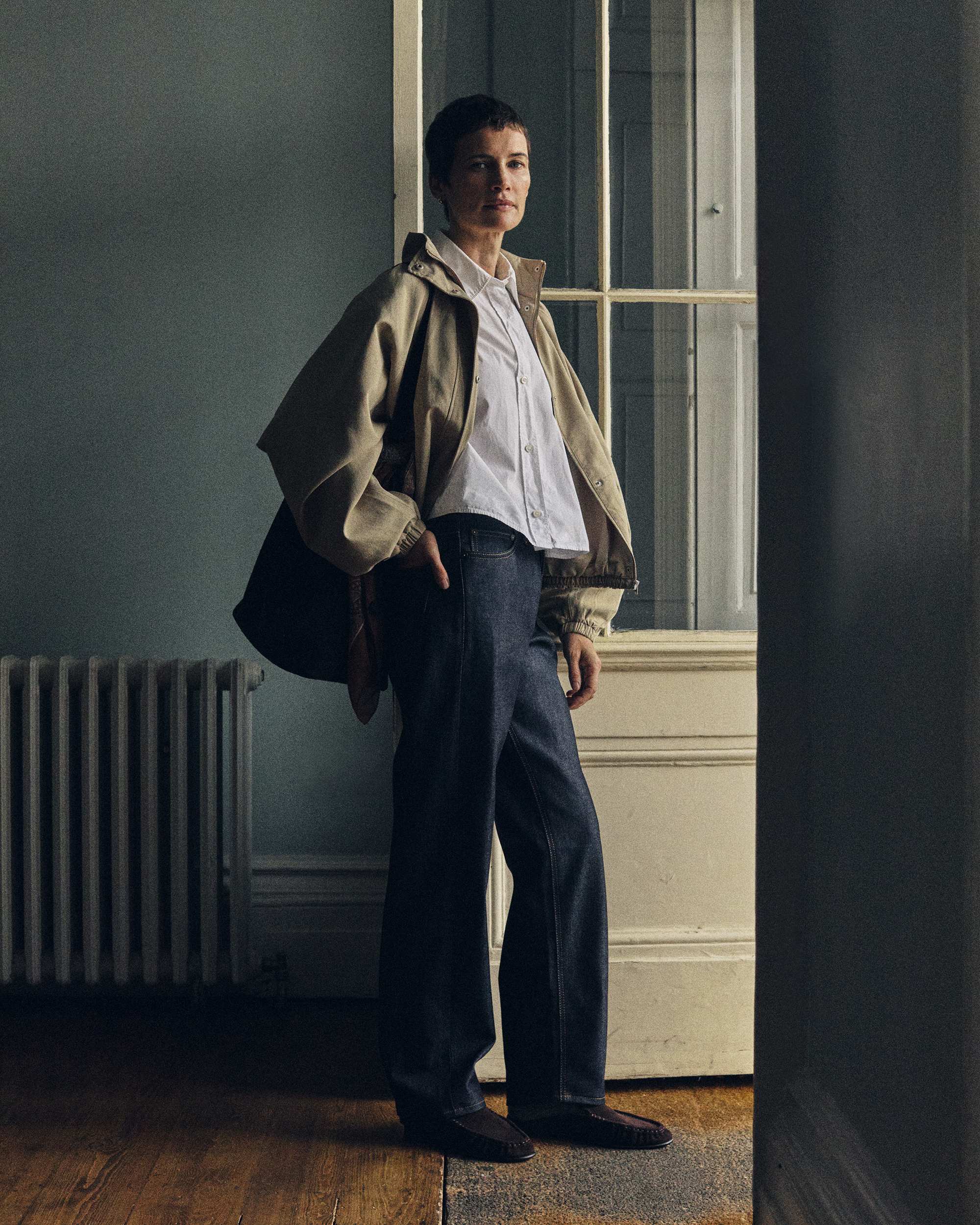Woman stands in doorway wearing Super Zero selvedge Hiut jeans, white shirt, and cream jacket. Also holding a bag.