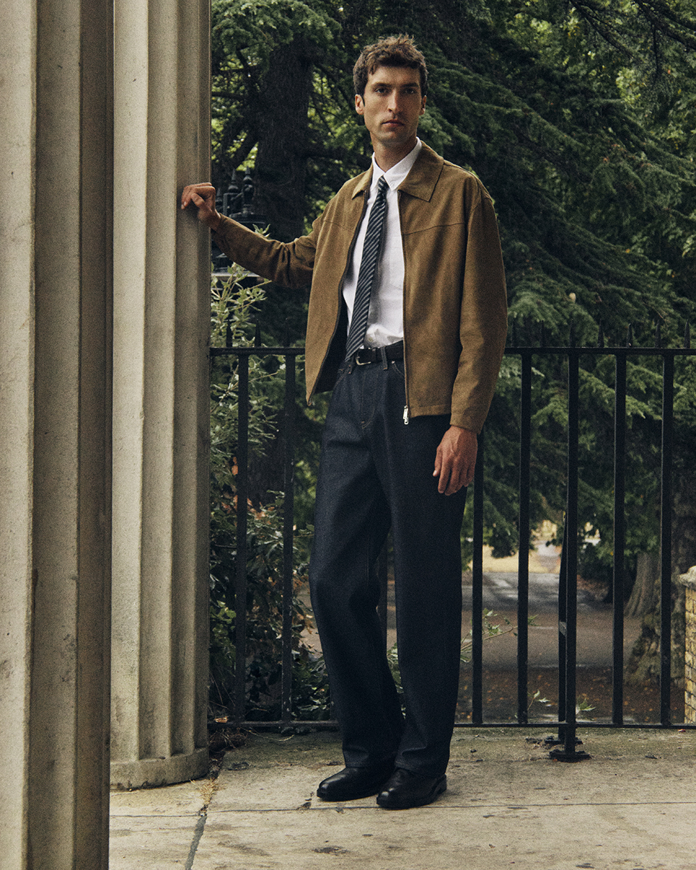 Man wearing dark selvedge jeans and a brown jacket beside a stone doorway.