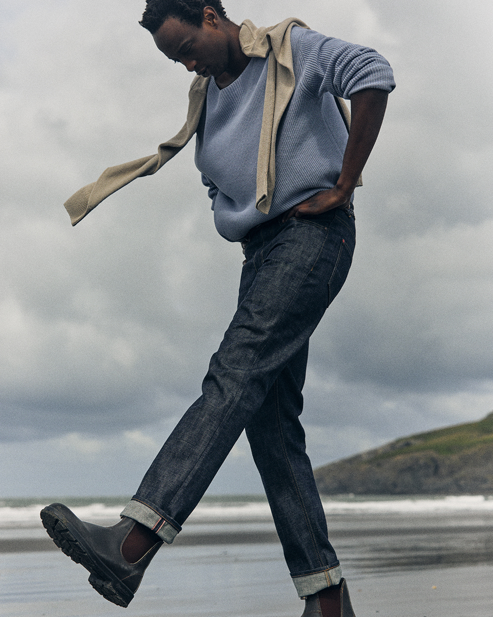 Photo of man standing on beach in selvedge denim jeans, brown boots, blue jumper and cream jumper over his shoulders.