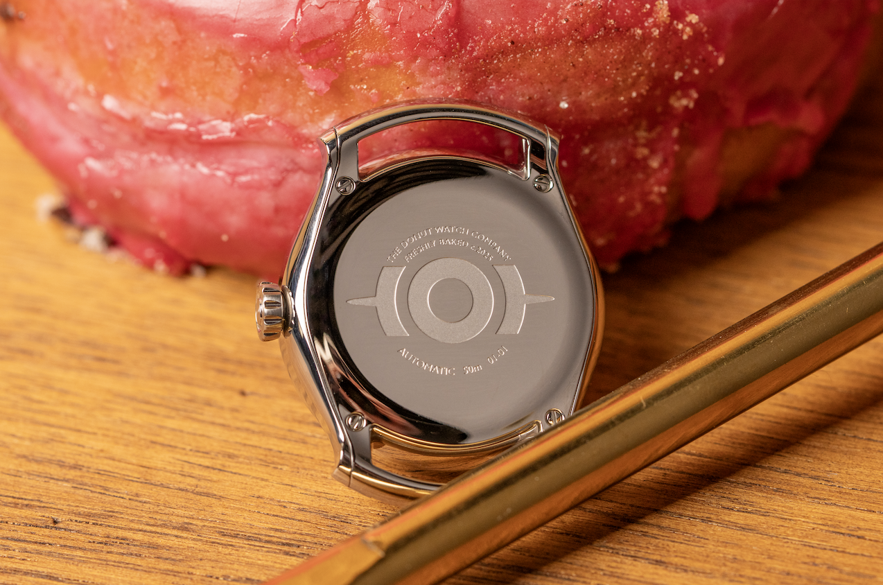 A polished caseback engraved 'The Donut Watch Company, Freshly Baked', resting against a glazed donut.
