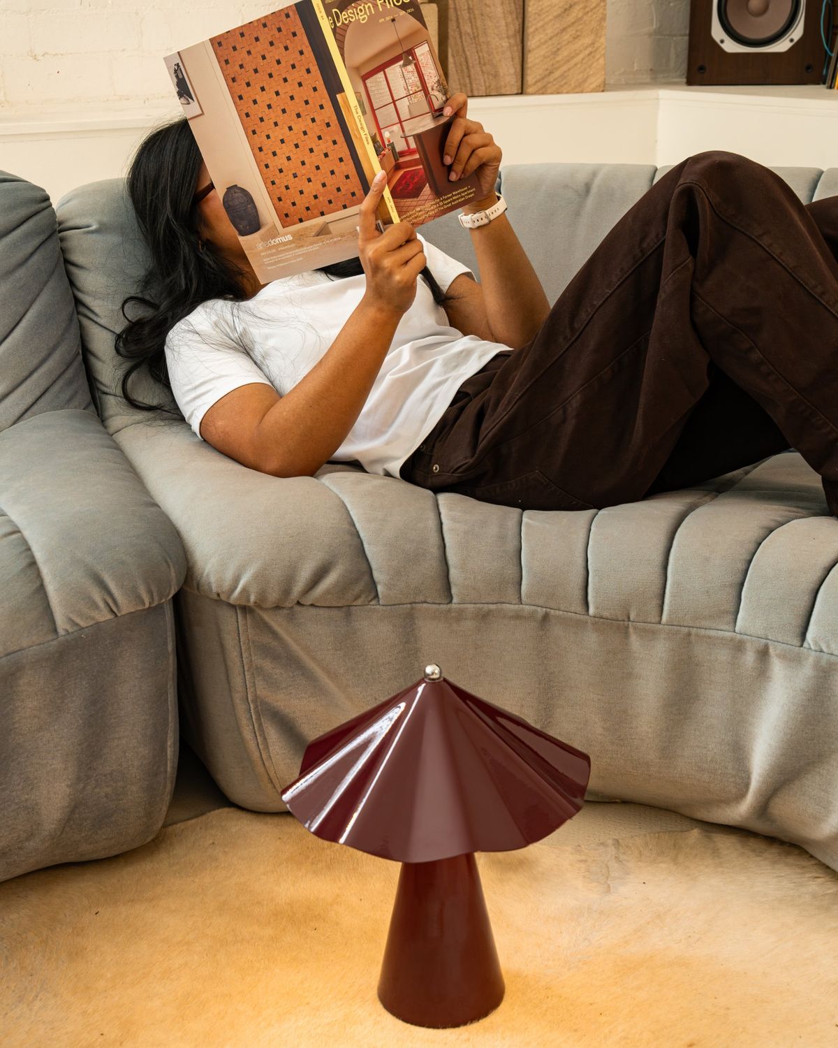 A Burgundy Robyn lamp on the floor beside someone reading on a sofa