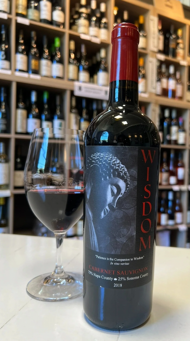 Bottle of B. Wise Wisdom 2018 Cabernet Sauvignon beside a glass of red wine, displayed on a white countertop with shelves of wine bottles in the background. Fearrington Village.