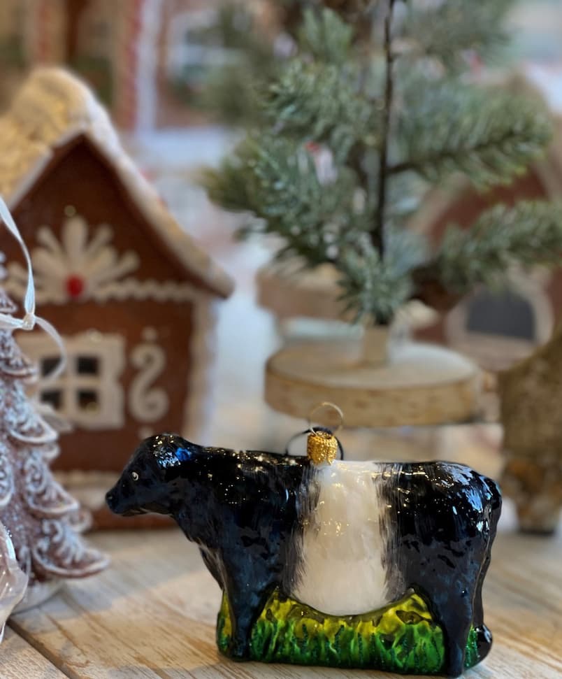 Glossy black-and-white Belted Galloway cow Christmas ornament displayed on a rustic wooden table alongside a miniature evergreen tree, gingerbread house, and festive holiday decorations. Fearrington Village.