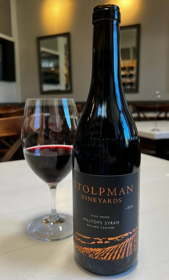 Bottle of Stolpman Vineyards 2018 Hilltops Syrah from Ballard Canyon, displayed beside a glass of red wine on a white table in a warmly lit dining room. Fearrington Village.