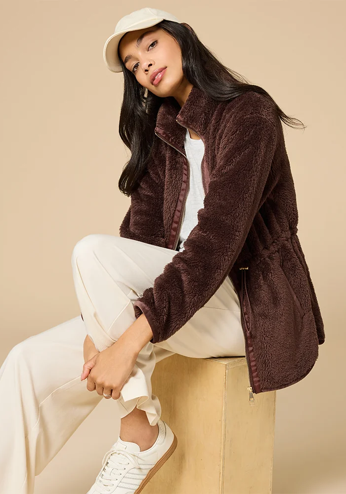 Model wearing a rich chocolate-brown fleece zip-up jacket, cream trousers, white sneakers, and a beige baseball cap, seated against a neutral studio backdrop. Fearrington Village.