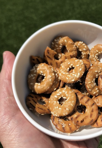 A white bowl filled with golden, ring-shaped protein pretzels, sprinkled with sesame seeds and savory seasonings, held outdoors in bright sunlight. Fearrington Village.