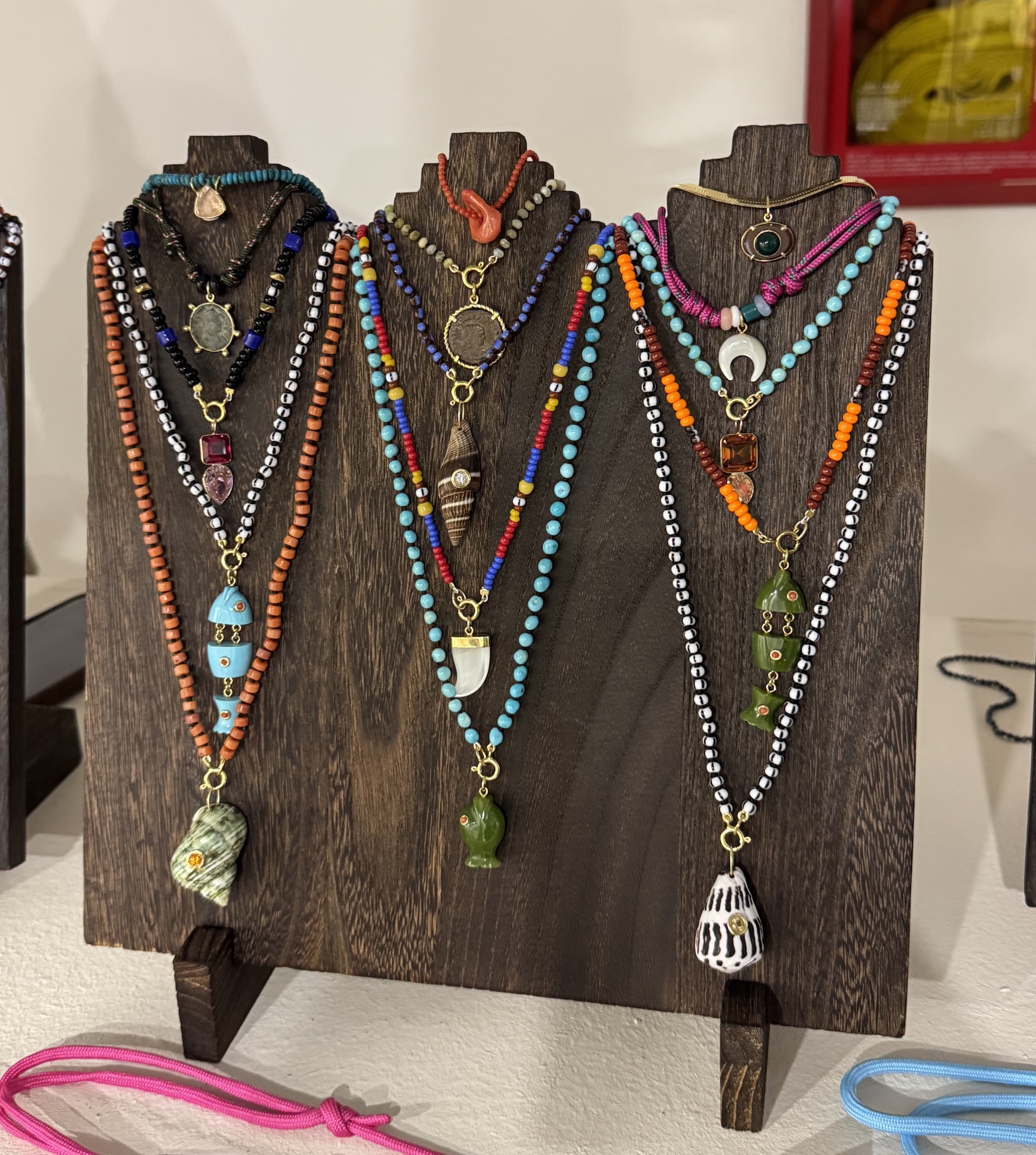 Colorful layered beaded necklaces with stone, shell, and sculptural pendants displayed on dark wood stands. Fearrington Village.