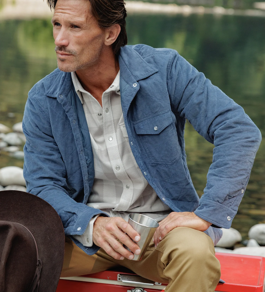 Man wearing a blue quilted corduroy shirt jacket over a light gray plaid shirt and khaki pants, seated by a river holding a stainless steel cup, with a brown cowboy hat beside him. Fearrington Village.