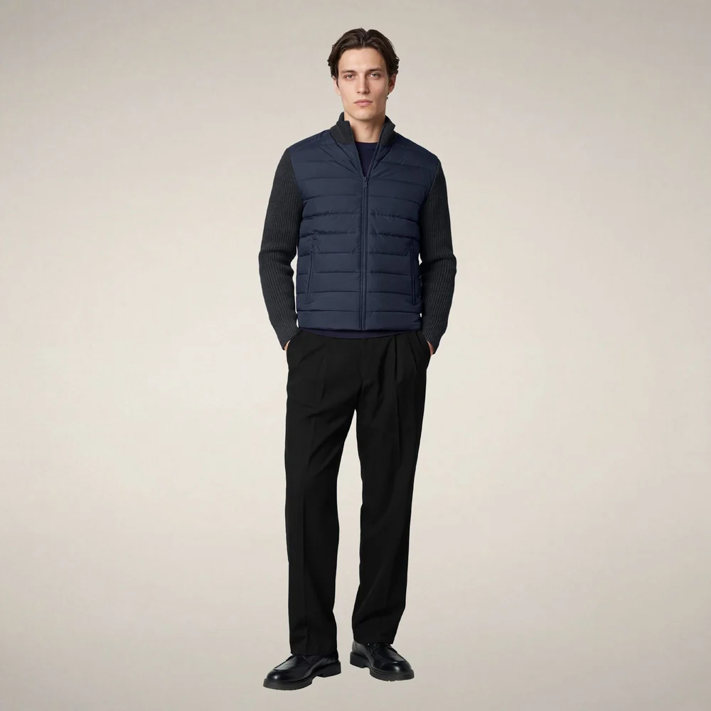 Model wearing a navy quilted zip-front jacket with ribbed knit sleeves, paired with black trousers and black leather shoes against a neutral beige background. Fearrington Village.