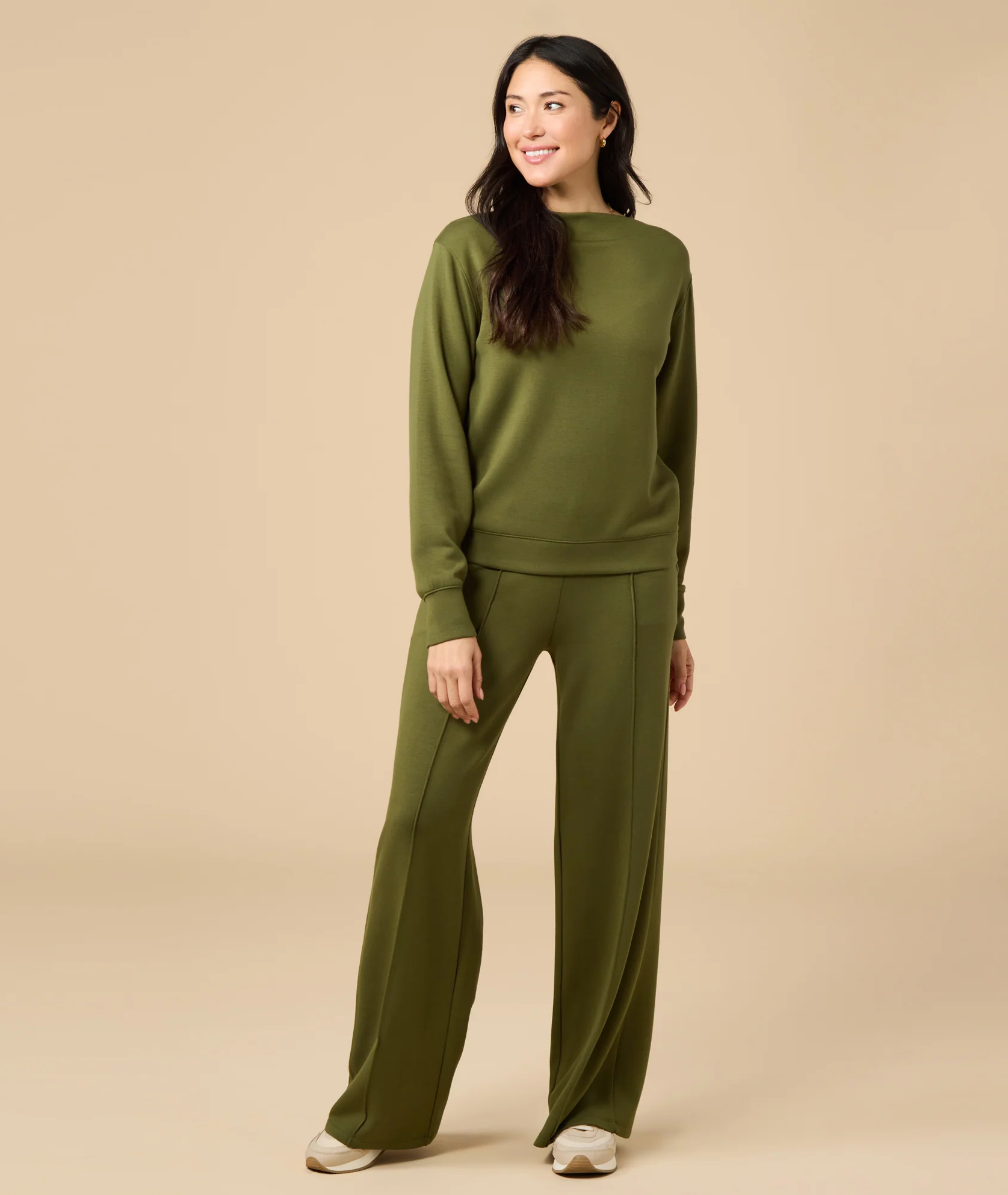 Model wearing an olive-green long-sleeve pullover with matching wide-leg pants and cream sneakers against a warm beige background. Fearrington Village.