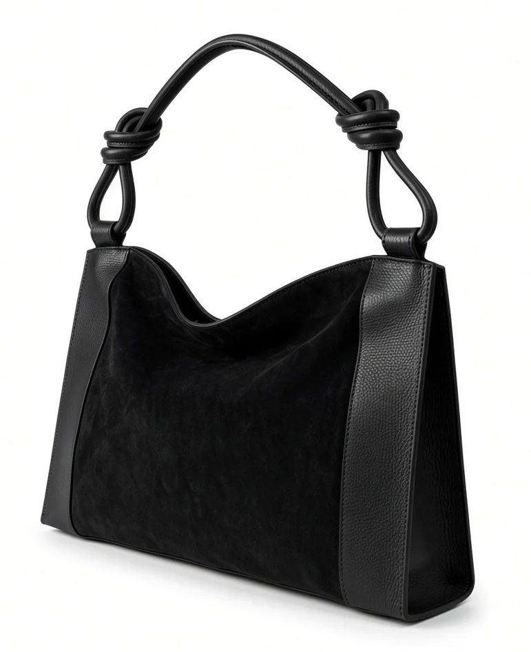 Black suede handbag with smooth leather side panels and a sculptural knotted leather handle, featuring a softly curved silhouette. Fearrington Village.