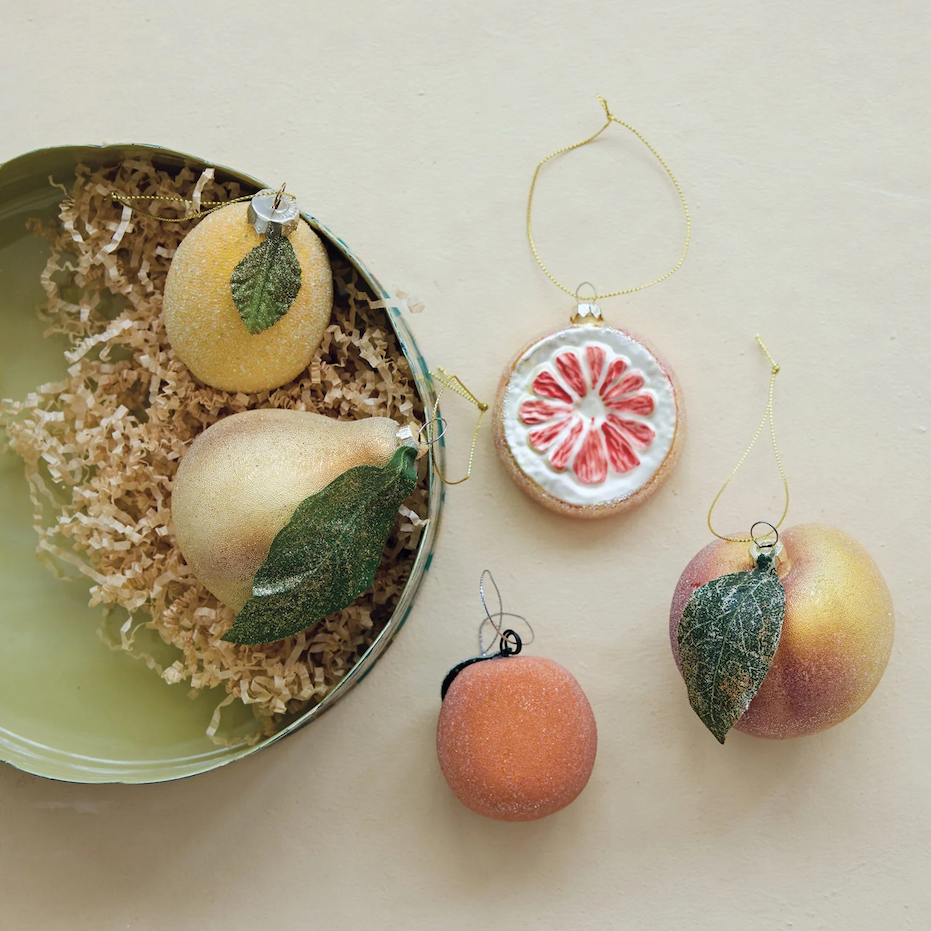 Assortment of glittering fruit-shaped Christmas ornaments, including lemons, a pear, a grapefruit slice, a peach, and an orange, displayed beside a pale green bowl filled with crinkle paper. Fearrington Village.