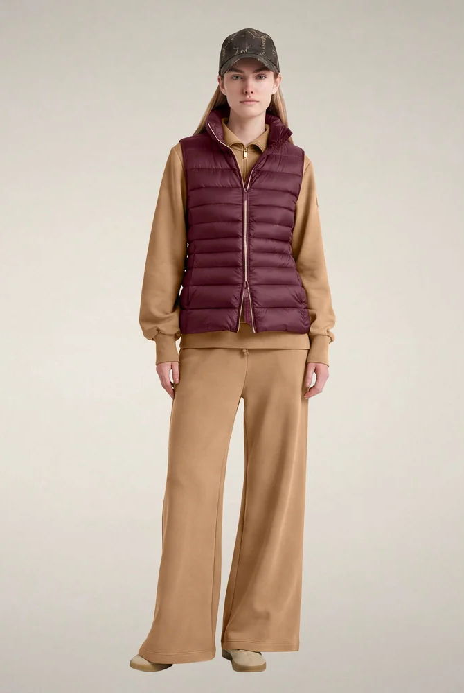 Model wearing a burgundy quilted puffer vest over a camel-colored sweatshirt and matching wide-leg trousers, finished with a dark patterned baseball cap and beige shoes against a neutral studio background. Fearrington Village.