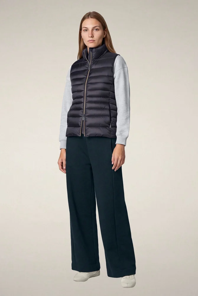 Model wearing a dark charcoal horizontal-quilted puffer vest over a light gray sweatshirt, paired with navy wide-leg trousers and white sneakers against a neutral studio background. Fearrington Village.