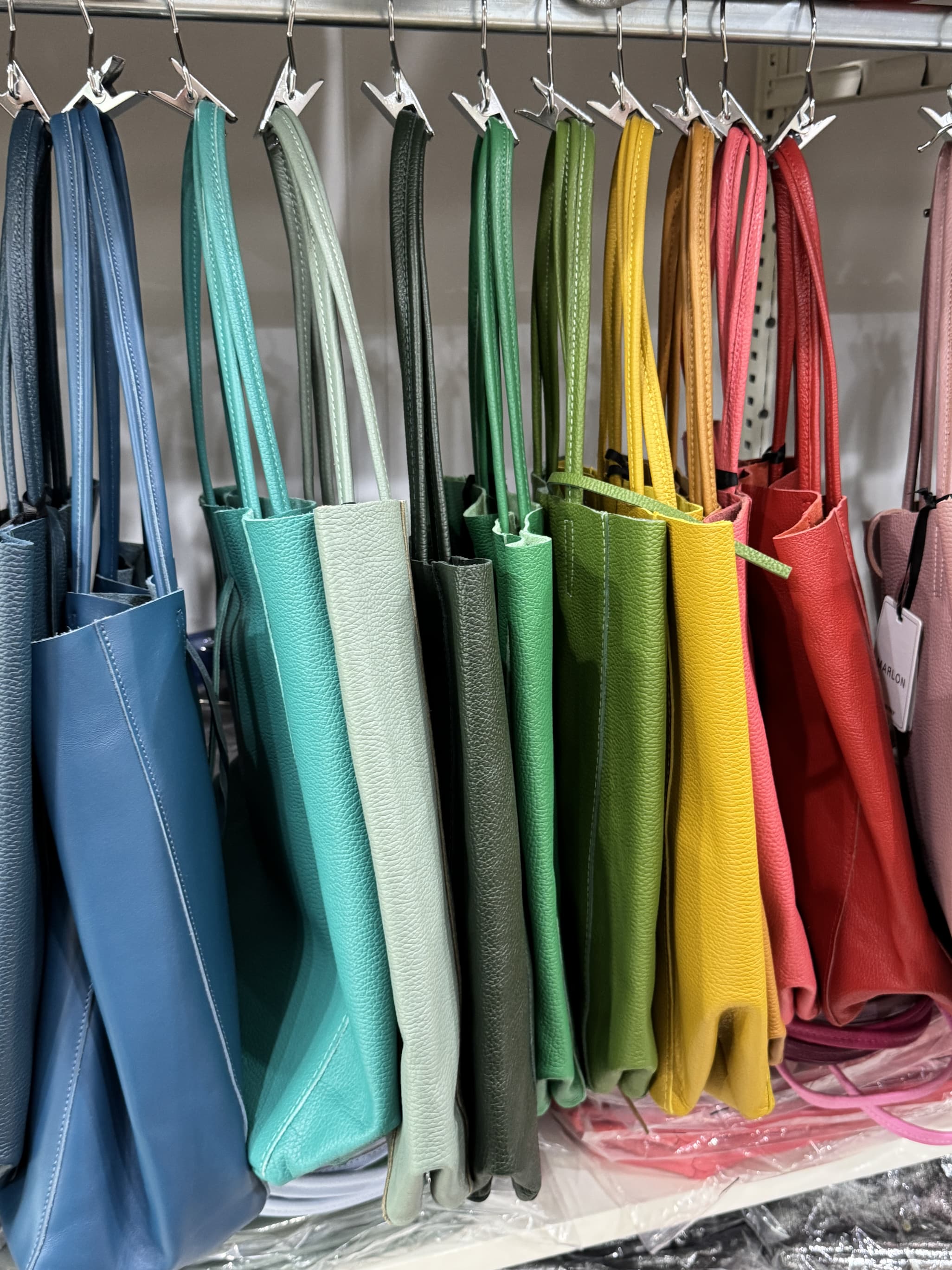 Colorful leather tote bags in shades of blue, turquoise, green, yellow, orange, red, and pink hanging on a rack. Fearrington Village.