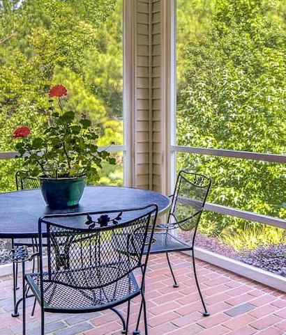 Screened porch with black metal patio furniture and a potted red geranium overlooking lush green trees and gardens. Fearrington Village.