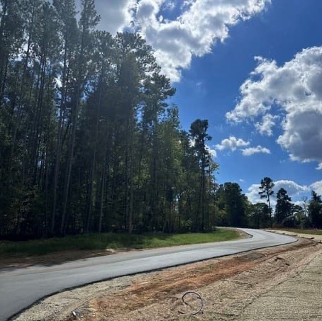 Newly paved road curves through a wooded landscape beneath a bright blue sky with billowing clouds. Fearrington Village.