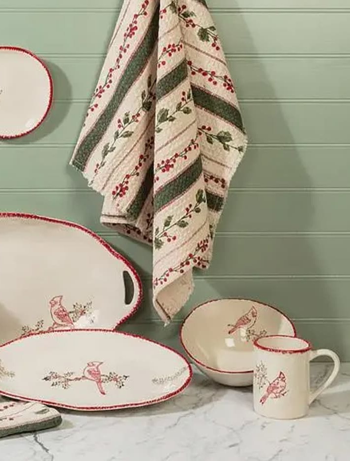 Festive ceramic serveware featuring red cardinal illustrations and scalloped red trim, displayed with coordinating mugs, pitchers, bowls, and holiday tea towels against a sage green backdrop. Fearrington Village.