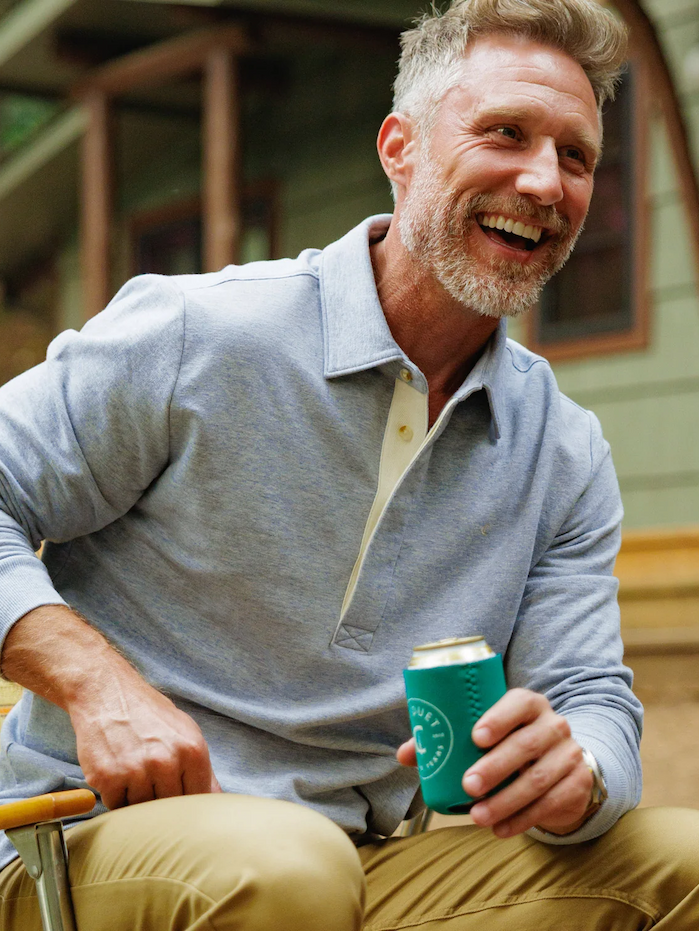 Smiling man with short gray hair and a beard wearing a light blue long-sleeve polo and khaki pants, seated outdoors and holding a beverage in a green koozie. Fearrington Village.