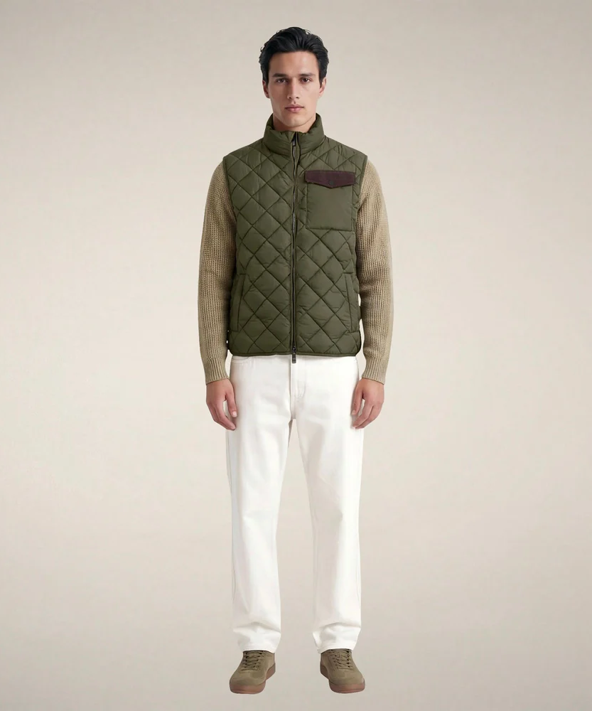 Model wearing an olive green diamond-quilted zip-front vest over a beige knit sweater, paired with ivory trousers and taupe suede shoes against a neutral background. Fearrington Village.
