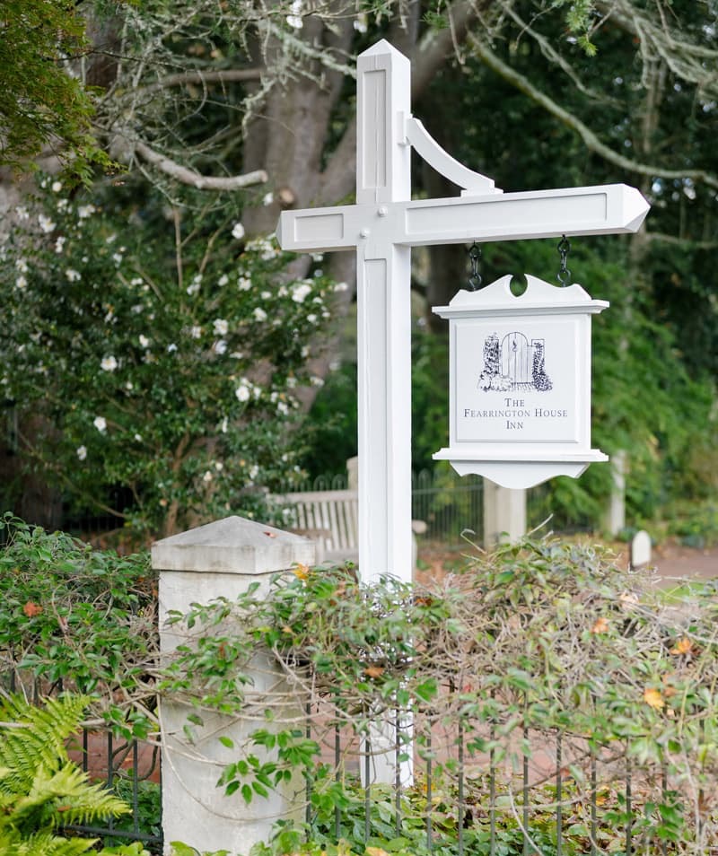White wooden sign for The Fearrington House Inn hanging from a traditional post, surrounded by lush greenery, flowering shrubs, and a garden fence. Fearrington Village.
