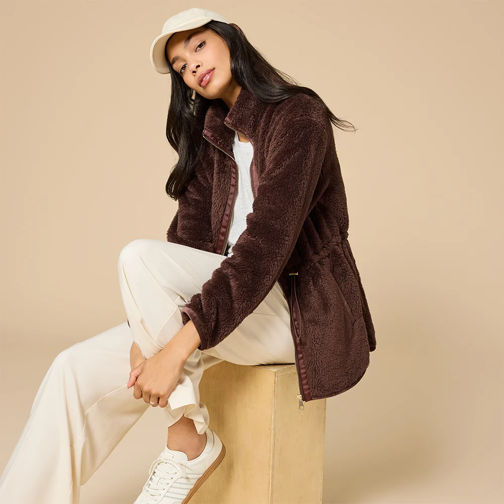 Model wearing a chocolate-brown plush fleece zip-up jacket with a high collar, cream pants, white