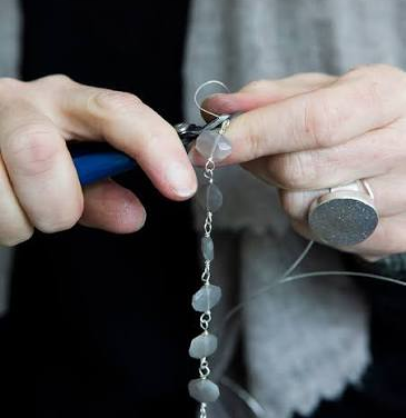 Close-up of a jewelry artisan using pliers to handcraft a necklace with pale blue-gray beads and delicate silver links. Fearrington Village.