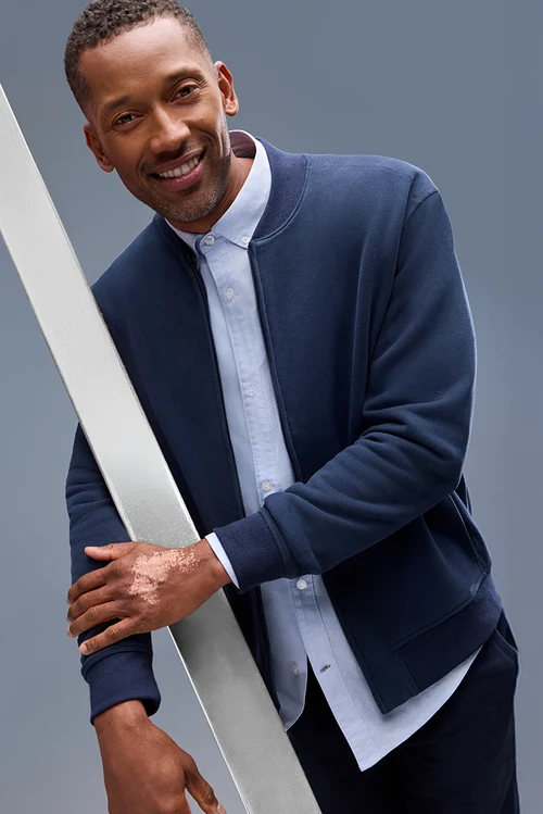 Smiling man wearing a navy jacket over a light blue button-down shirt against a blue-gray background. Fearrington Village.