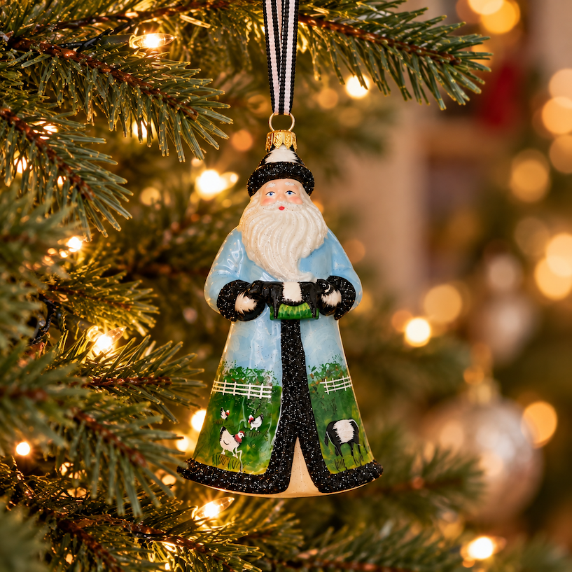 Hand-painted Santa Christmas ornament in a pale blue coat featuring Belted Galloway cows and chickens, hanging from a green Christmas tree with warm twinkling lights and a softly blurred background. Fearrington Village.