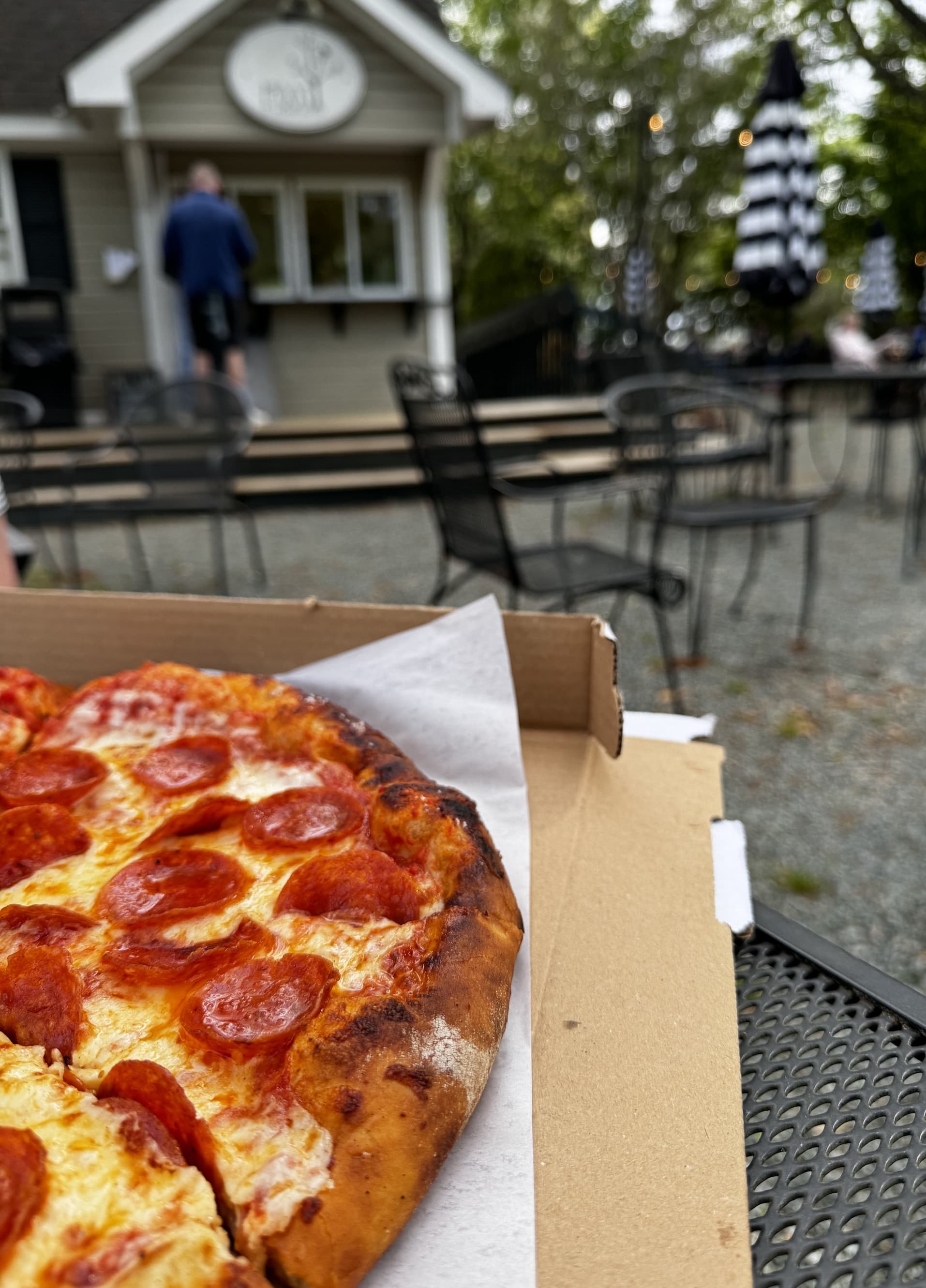 Close-up of a pepperoni pizza in an open box on a metal patio table, with a blurred outdoor café setting in the background. Behind it, a small food stand and scattered tables with black-and-white striped umbrellas sit among trees, suggesting a casual, relaxed dining area.