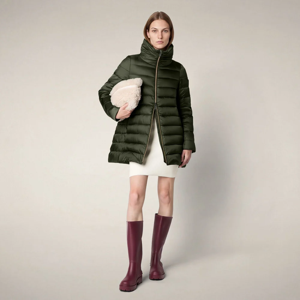 Model wearing a forest green, knee-length quilted puffer coat with a high collar, paired with a cream mini dress, burgundy knee-high boots, and a plush ivory clutch against a neutral studio background. Fearrington Village.