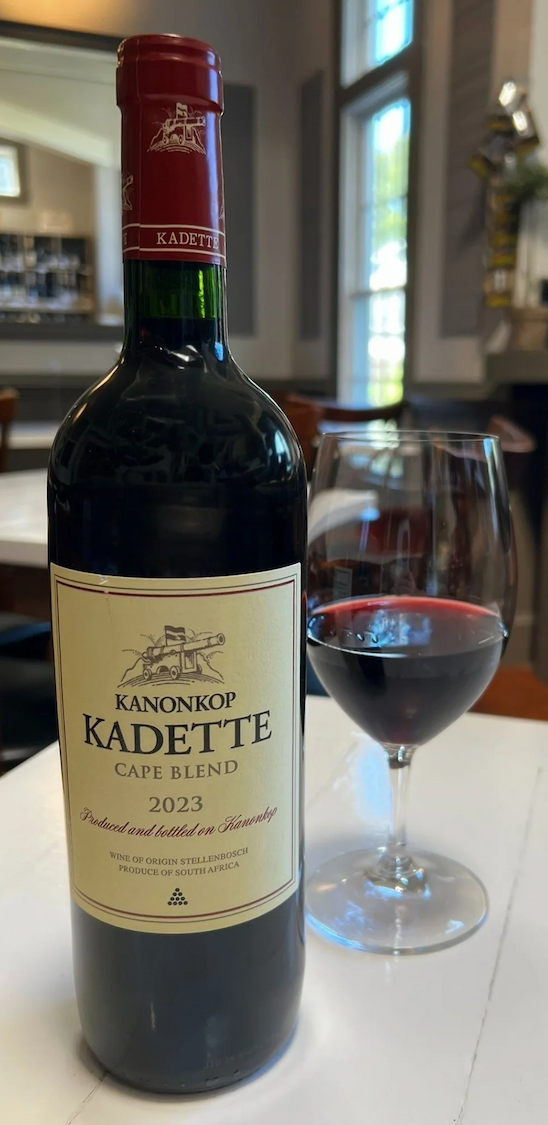 Bottle of Kanonkop Kadette Cape Blend 2023 beside a glass of red wine on a white table in a softly lit wine bar. Fearrington Village.