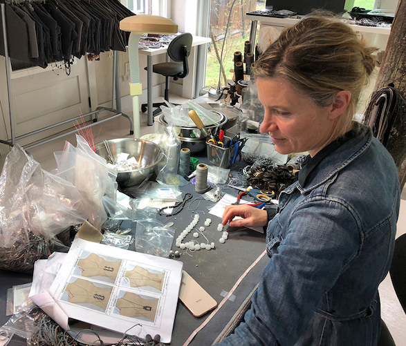 Jewelry designer in a denim jacket arranging beads and materials at a worktable filled with tools, sketches, and supplies in her studio. Fearrington Village.