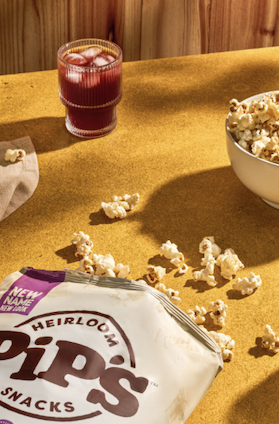 Bag of Pipcorn heirloom popcorn surrounded by scattered popcorn kernels, with a bowl of popcorn and a red drink on a mustard-yellow tabletop. Fearrington Village.