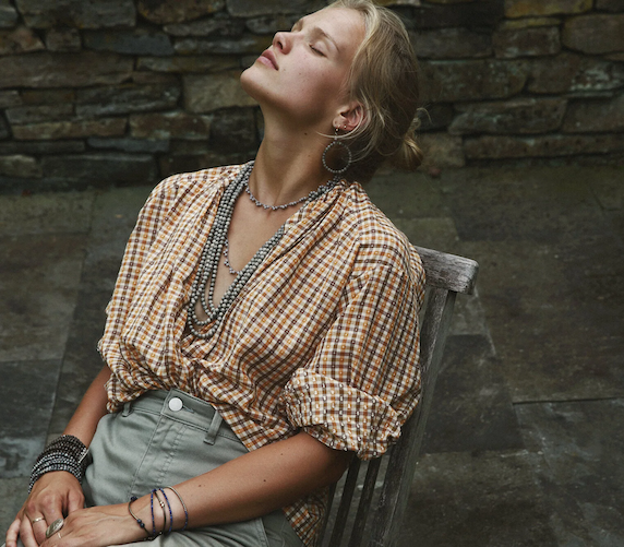 Woman wearing a mustard and white gingham blouse with puff sleeves, sage green trousers, and layered beaded jewelry, relaxing in a wooden chair against a rustic stone wall. Fearrington Village.