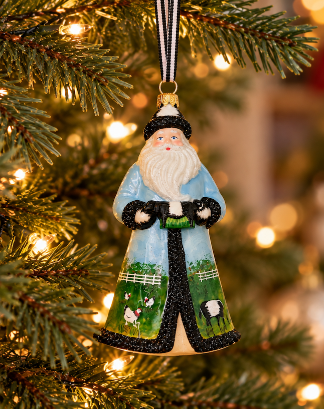 Hand-painted Santa Claus ornament in a blue coat decorated with farm animals and green pastures, hanging from a softly lit Christmas tree. Fearrington Village.