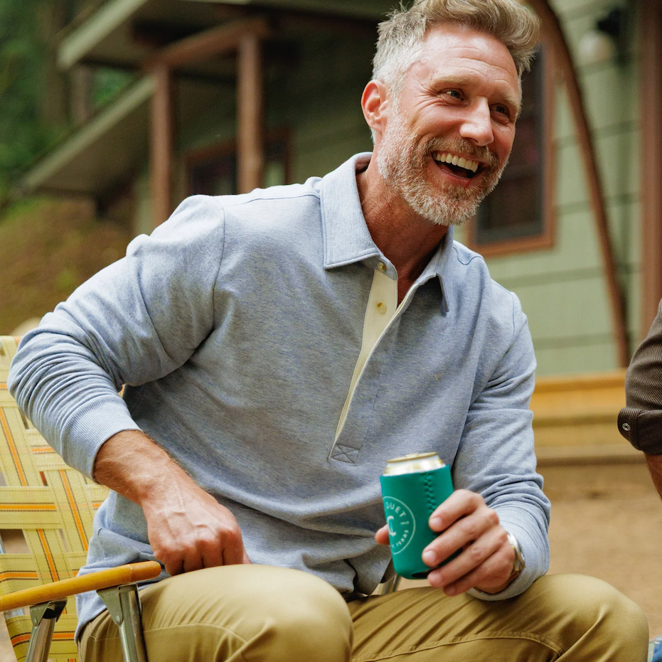 Smiling man wearing a light blue long-sleeve polo shirt and khaki pants, seated outdoors in a folding chair and holding a beverage can in a green koozie. Fearrington Village.