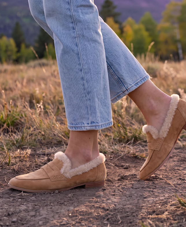 Close-up of tan suede loafers lined with soft cream faux fur, paired with light-wash jeans and styled outdoors on a rustic path with autumn foliage in the background. Fearrington Village.