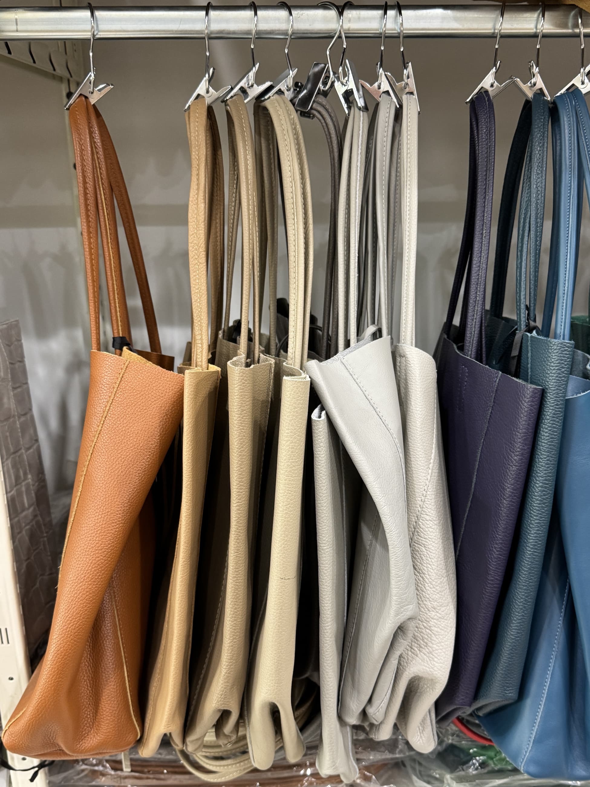 Leather tote bags in warm tan, neutral cream and gray, deep purple, and shades of blue hanging on a rack. Fearrington Village.