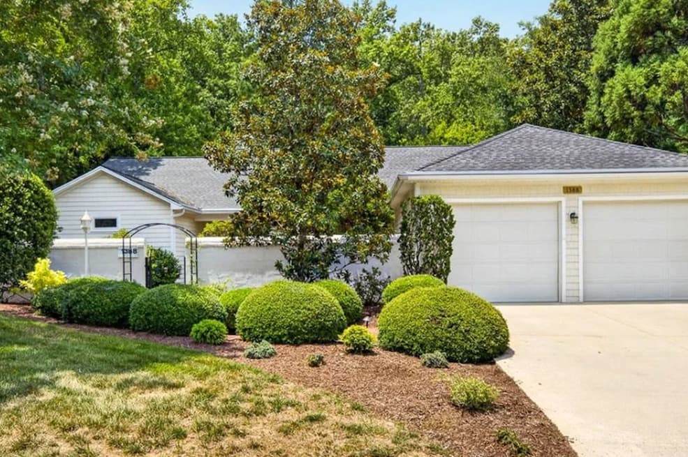 White cottage with a two-car garage, manicured shrubs, and mature trees surrounding the landscaped front yard. Fearrington Village.