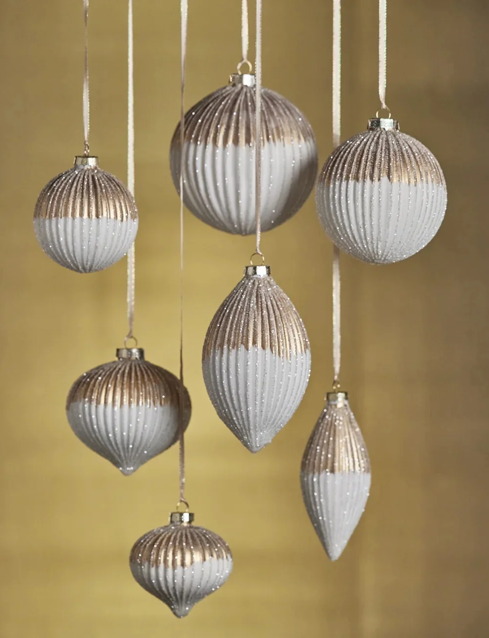 Collection of elegant ribbed glass Christmas ornaments in assorted round, teardrop, and finial shapes, featuring shimmering champagne-gold tops and frosted white finishes, suspended against a warm golden background. Fearrington Village.