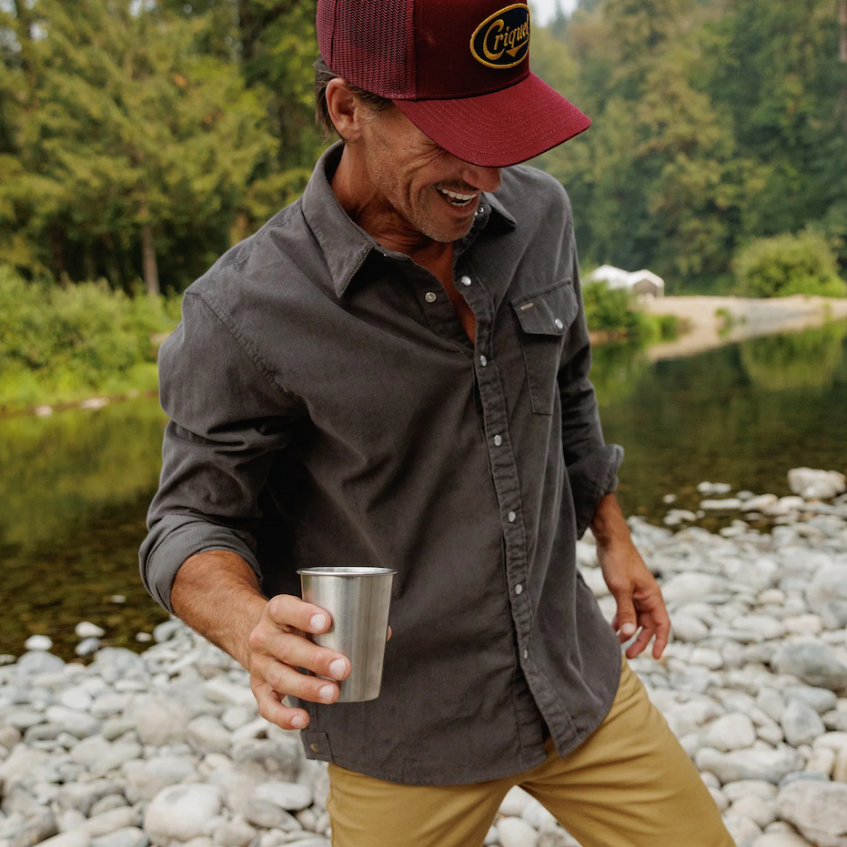 Man wearing a charcoal-gray button-down shirt, tan pants, and a burgundy Criquet baseball cap, holding a stainless steel cup beside a rocky river surrounded by greenery. Fearrington Village.