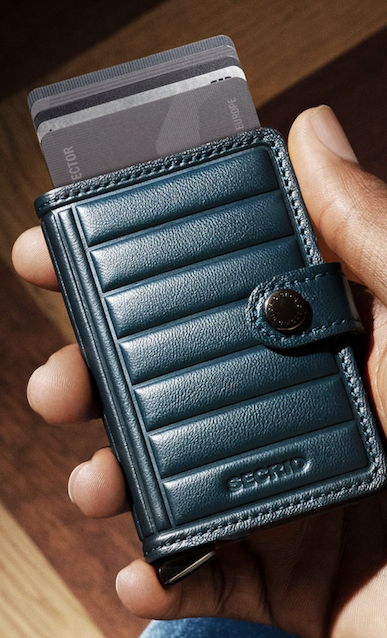 Close-up of a hand holding a compact navy blue Secrid leather wallet with a snap closure and several cards extending from its aluminum Cardprotector. Fearrington Village.