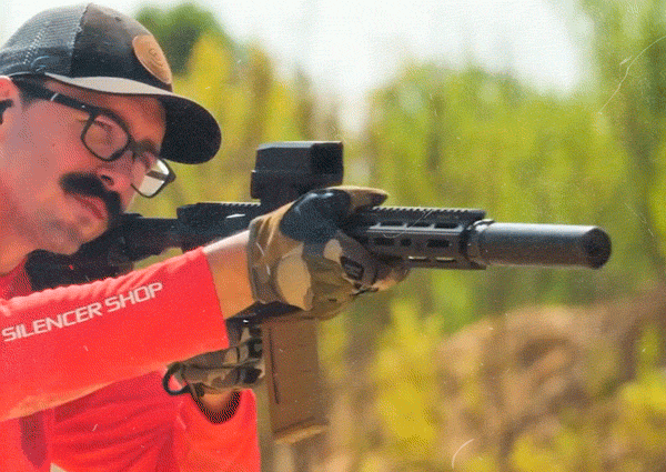 Person with suppressor-equipped rifle aiming in outdoor setting.