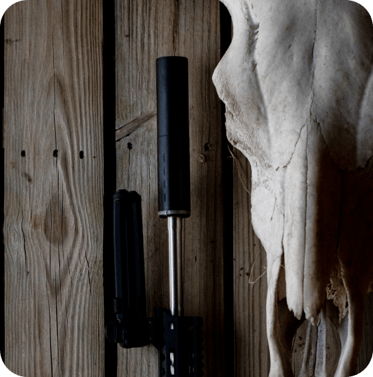Suppressor-mounted rifle leaning against a rustic wooden wall beside a deer skull, ready for a pre-season zero check.