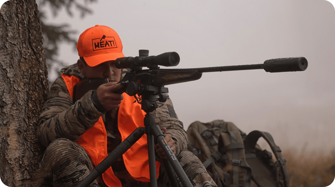 Outdoor hunting scene with person in orange cap and vest aiming rifle beside tree and gear.