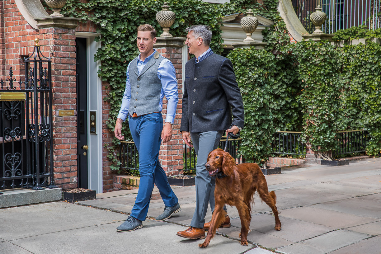 Two men in an Austrian style jacket and vest walking a dog along a leafy street
